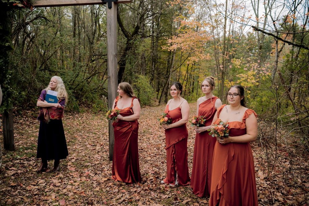 Four bridesmaids in rust-red gowns holding bouquets stand among autumn leaves in a wooded outdoor wedding ceremony setting.