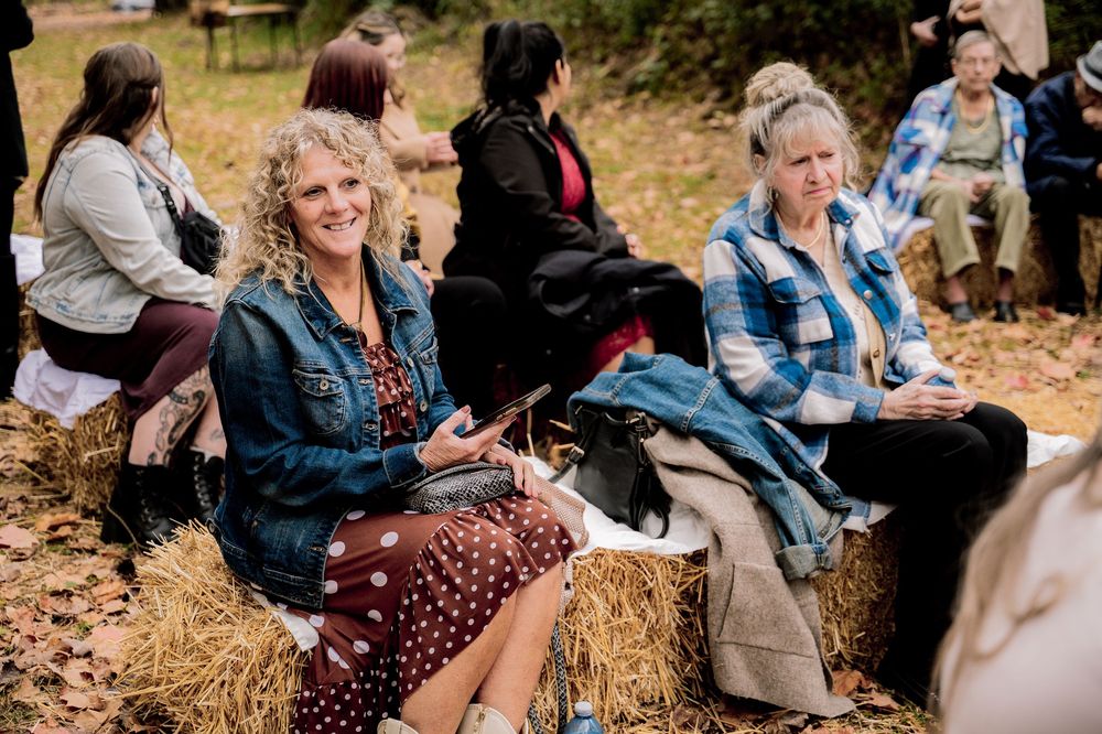 Women sitting on hay bales outdoors at a rustic autumn gathering, smiling and socializing in casual attire.