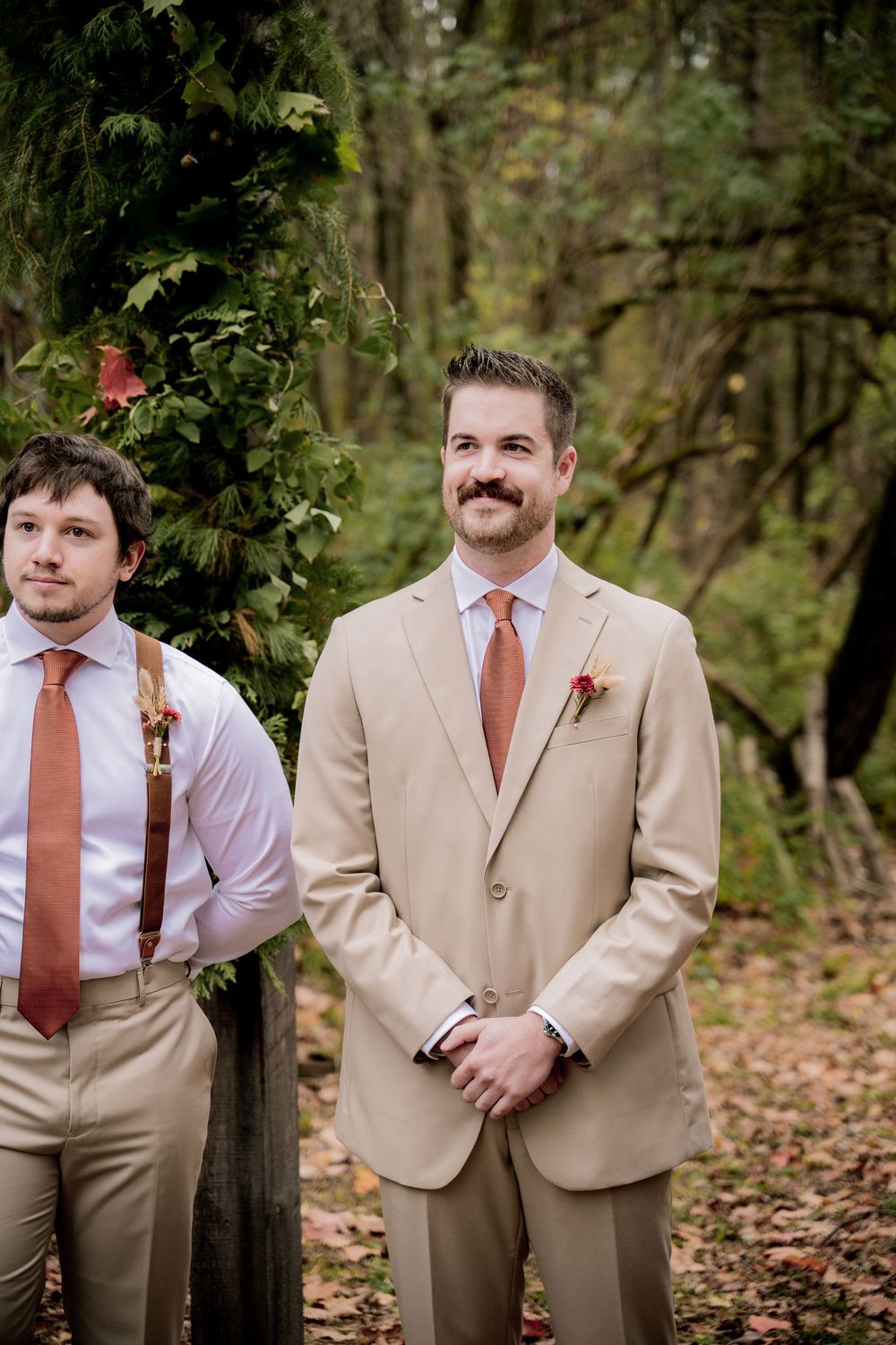 Groom in tan suit with orange tie and boutonniere stands outdoors at woodland wedding ceremony.