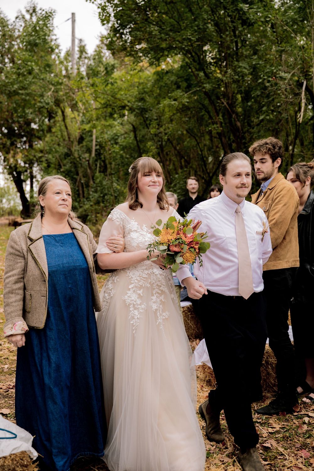 Bride in lace gown holding autumn bouquet walks with groom in pink shirt at outdoor fall wedding ceremony.