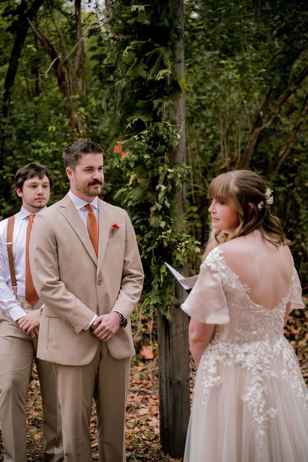 Groom in tan suit with orange tie smiles at bride reading vows in lace wedding dress at outdoor forest ceremony.