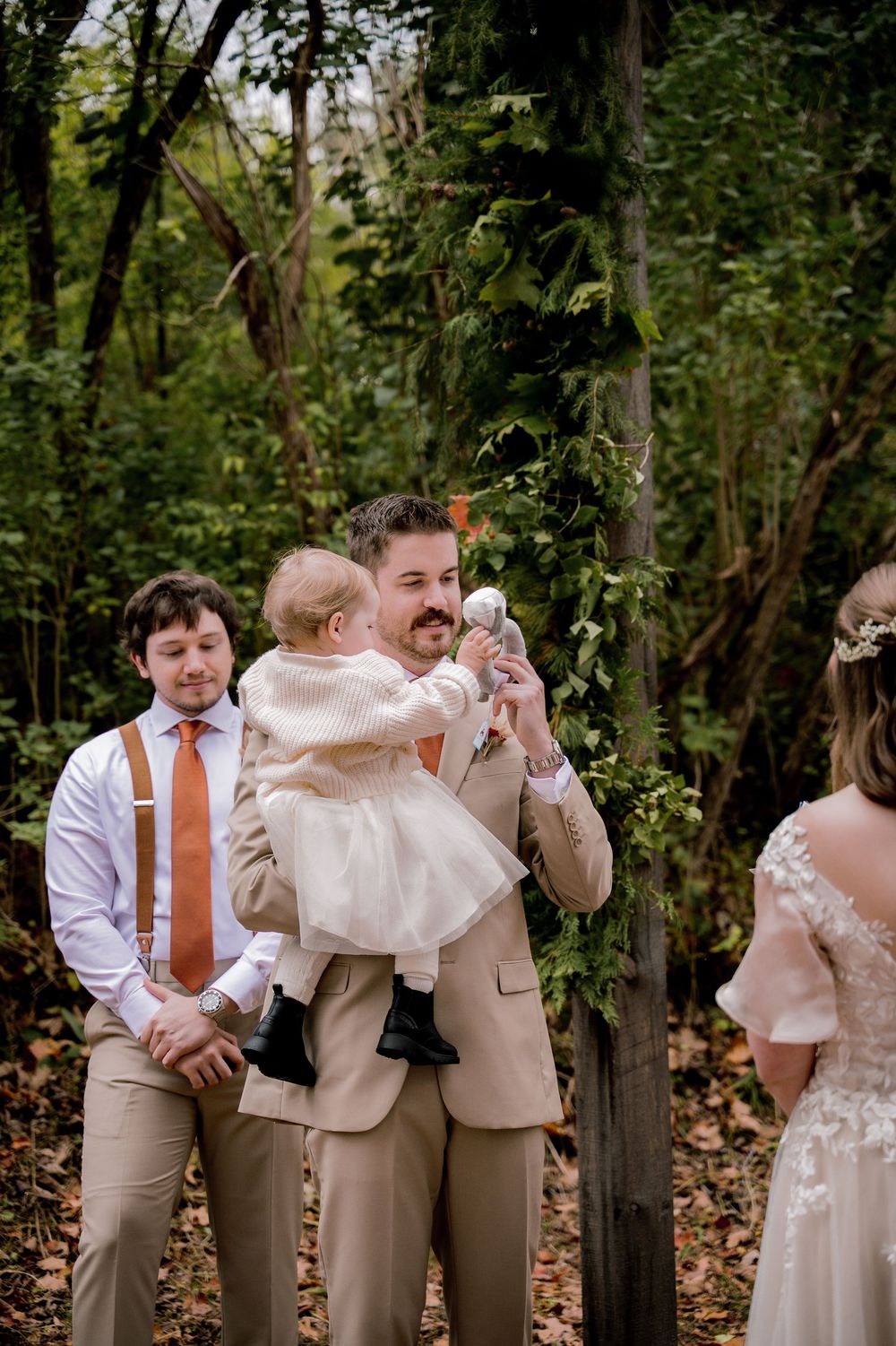 Groom in tan suit holds baby at outdoor woodland wedding ceremony while groomsman stands nearby in suspenders.