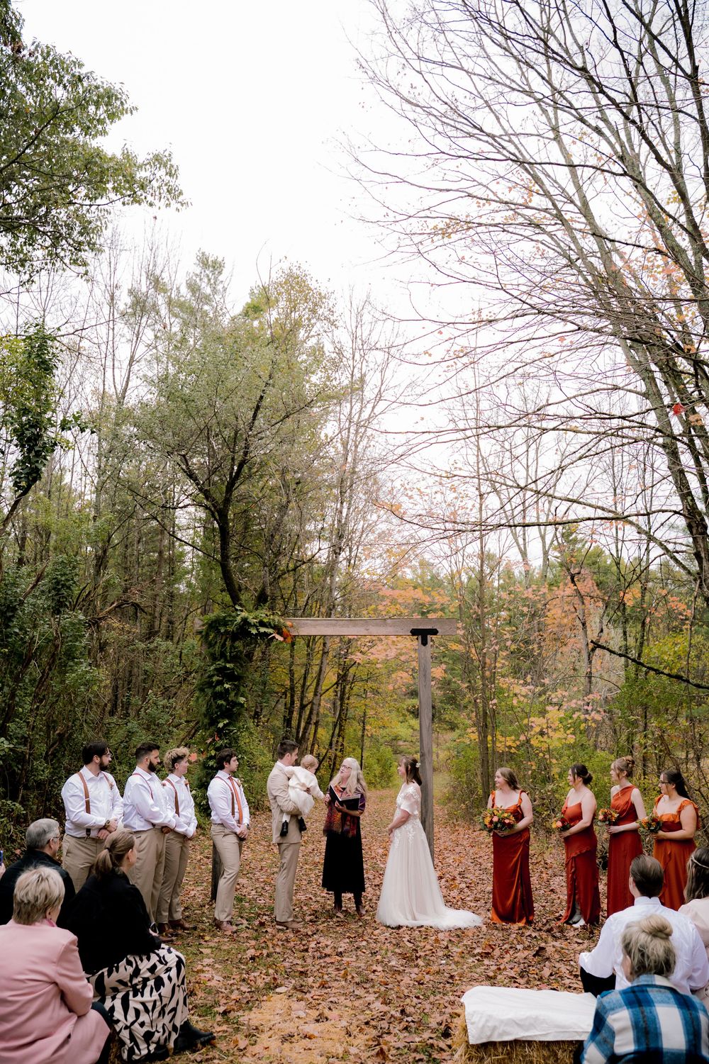 Outdoor fall wedding ceremony in forest with bridal party and guests among autumn leaves.