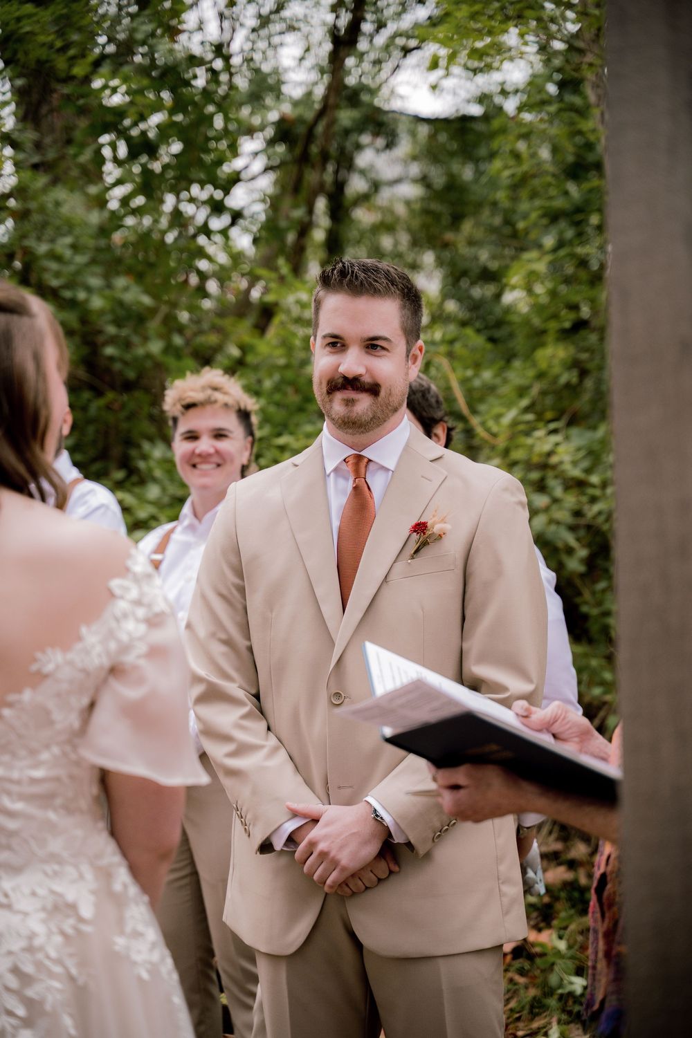 Groom in tan suit with orange tie smiling at bride during outdoor wedding ceremony with greenery backdrop.