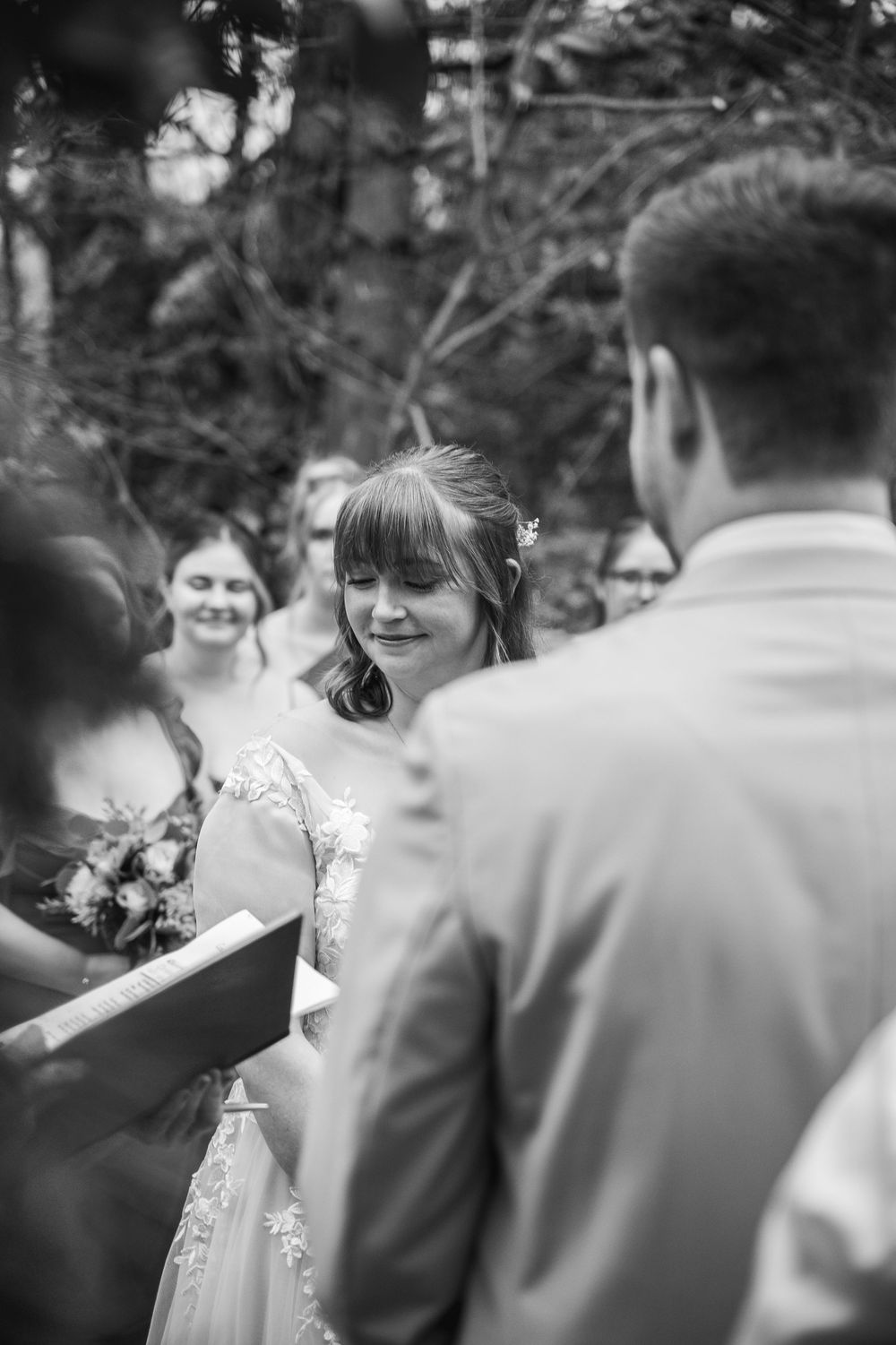 Black and white photo of a bride smiling during outdoor wedding ceremony with groom and guests in background.