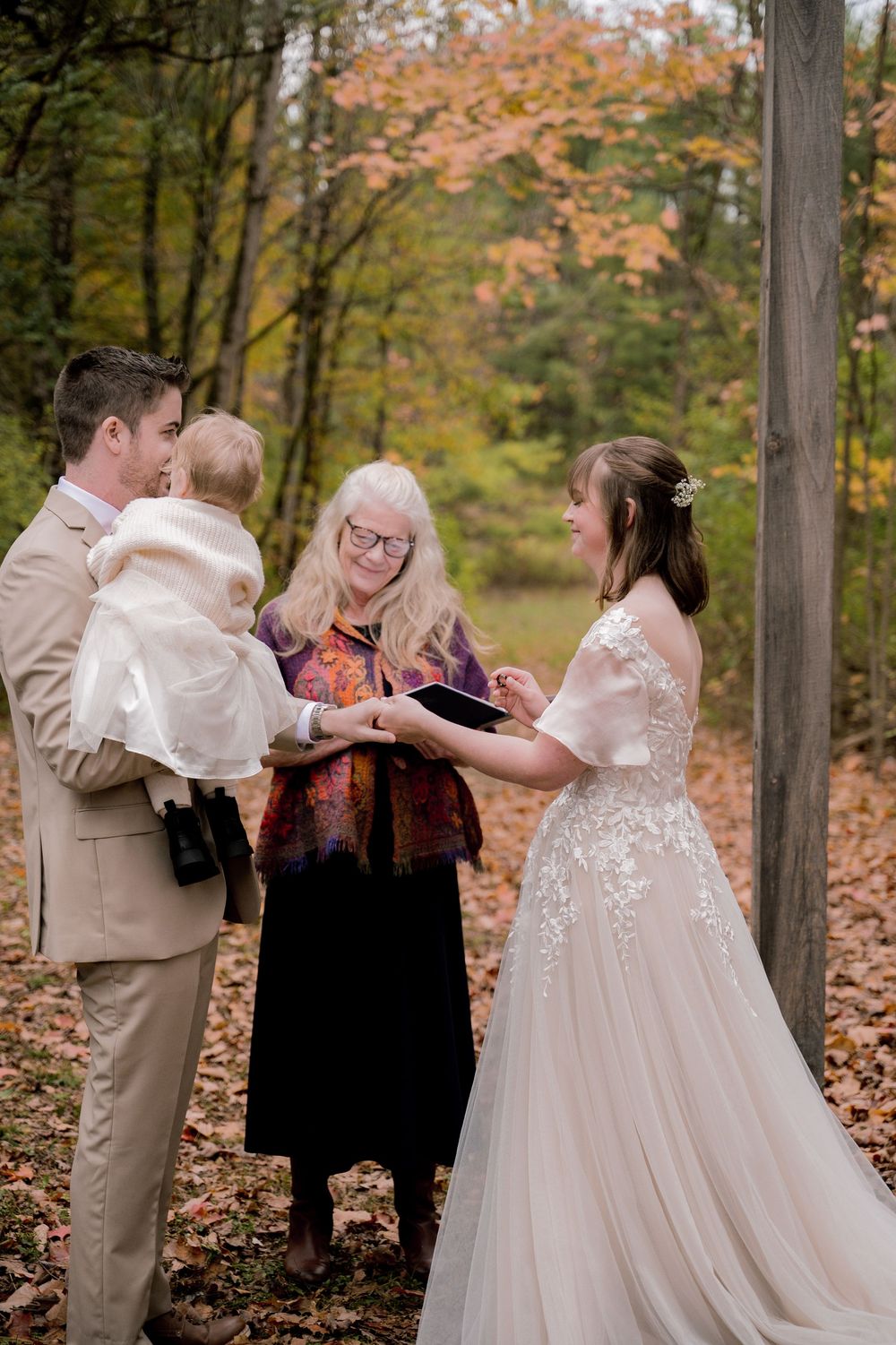 Bride and groom holding baby during outdoor fall wedding ceremony with officiant among autumn foliage.