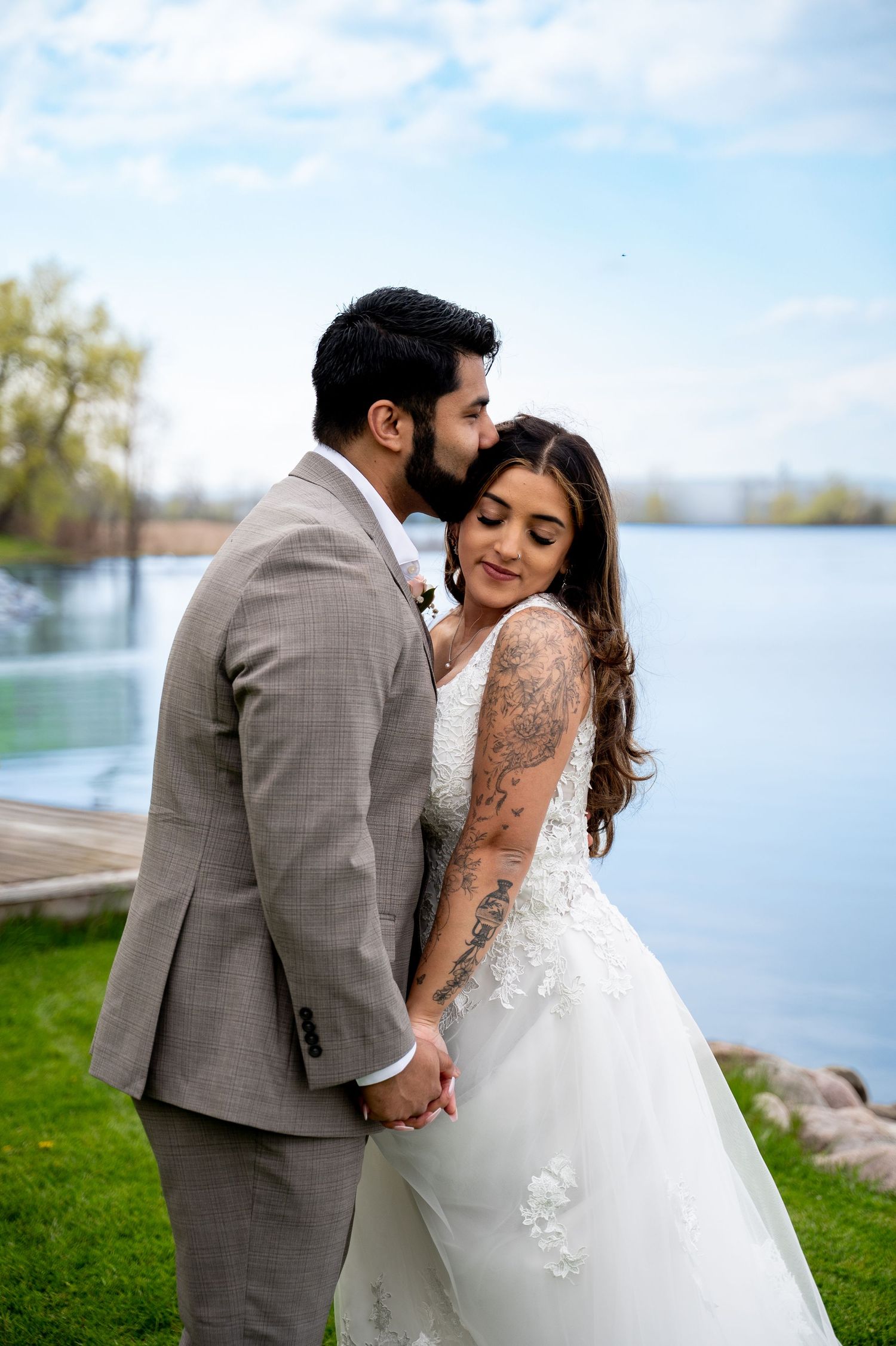 Groom in gray suit kisses bride in lace wedding dress by a lake, holding hands on their wedding day.