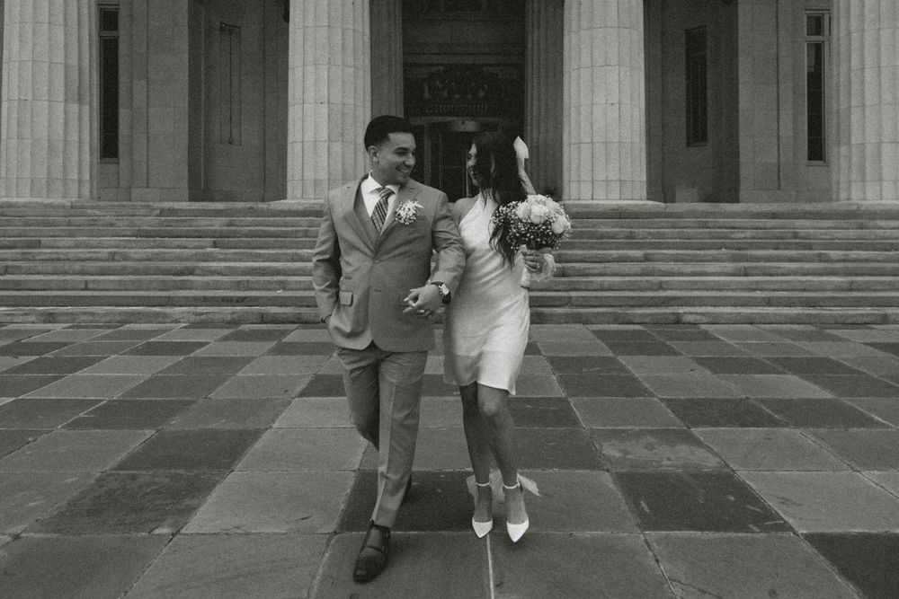 Black and white photo of a couple in wedding attire walking down steps outside a grand building.