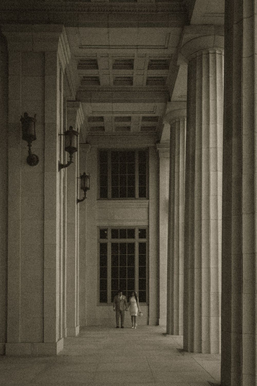 Couple standing in grand neoclassical building colonnade with tall stone pillars and ornate ceiling.