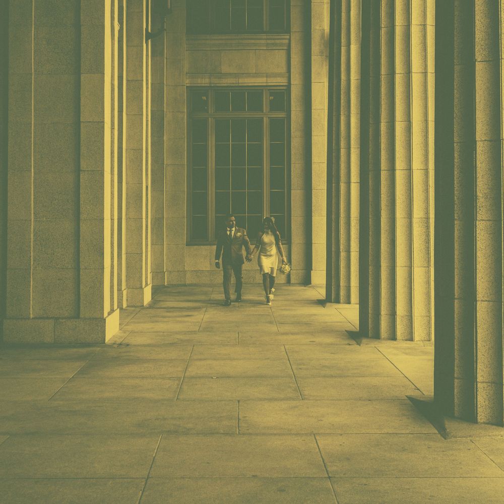 Two people walking through grand stone colonnade corridor with tall pillars and tiled floor in golden tones.