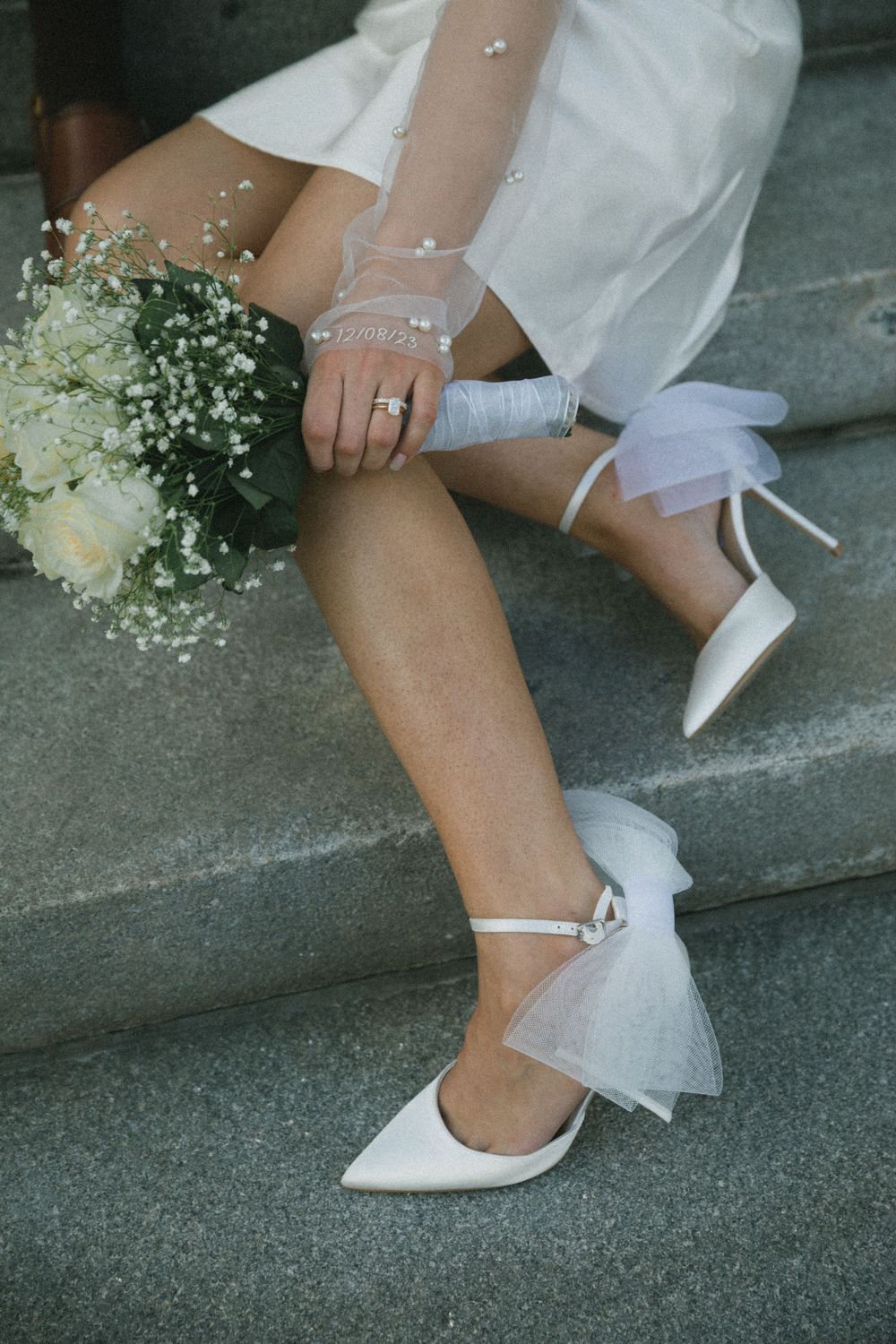 Bride sitting on stone steps wearing white pointed-toe heels with tulle bows, holding a white rose bouquet.