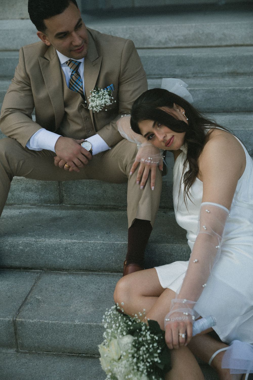 Bride and groom sitting on stone steps, she leans on his knee holding a bouquet in a white mini dress.