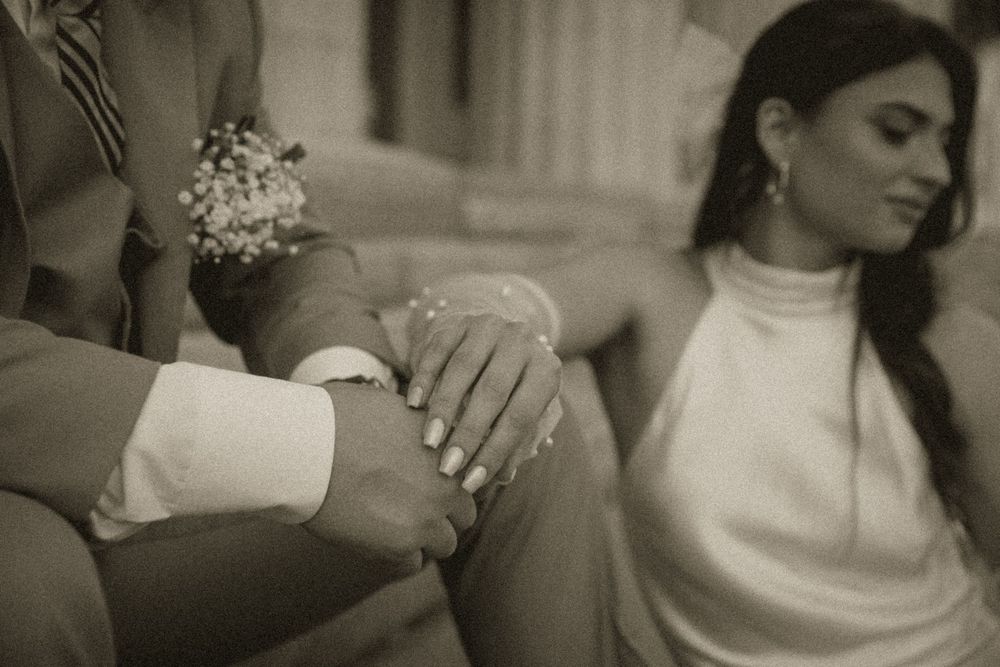 Black and white photo of a bride and groom holding hands, showcasing wedding rings, in elegant attire.