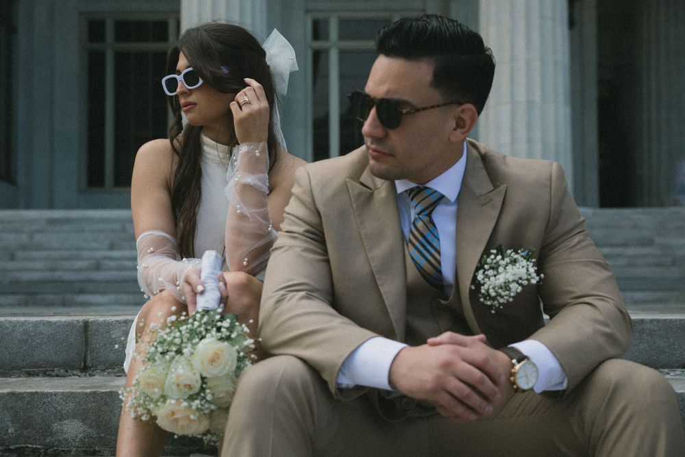 Elegant couple in wedding attire sitting on stone steps, groom in tan suit with boutonniere, bride holding bouquet.