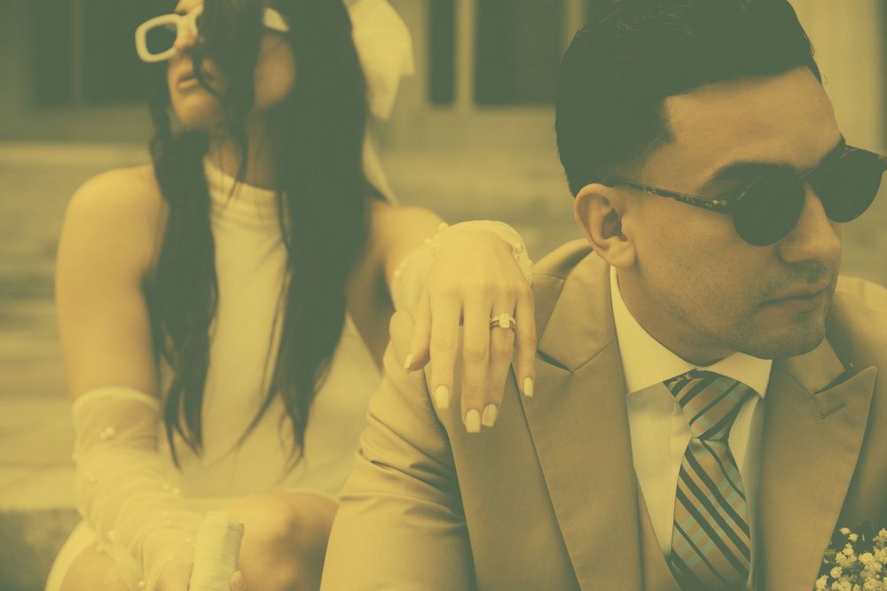 A bride and groom pose together outdoors, she in white dress with sunglasses, he in tan suit with striped tie.