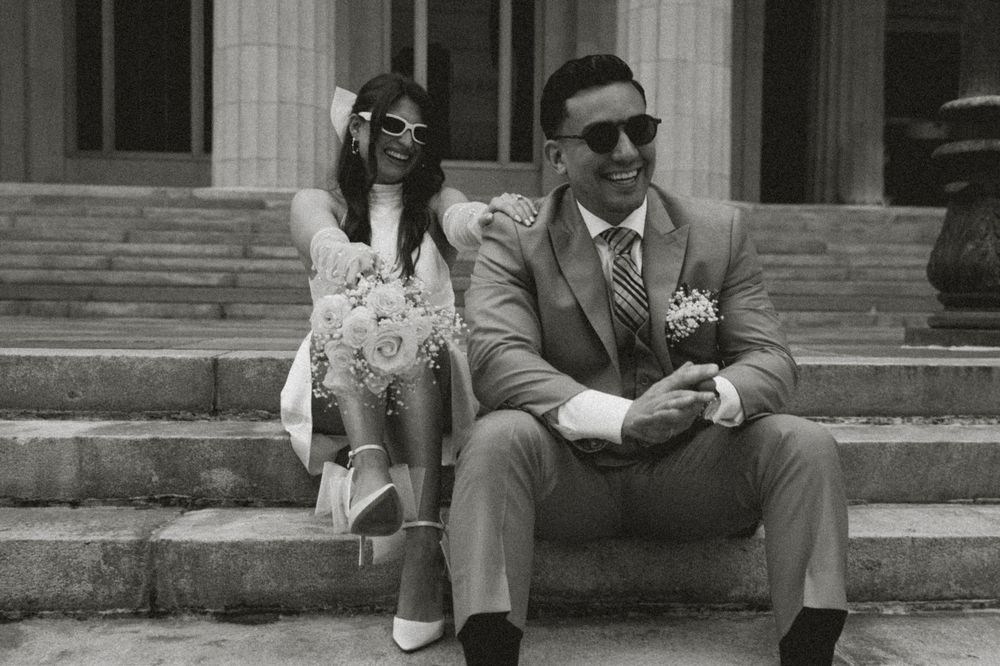 Black and white photo of a bride and groom sitting on steps, wearing sunglasses, smiling on their wedding day.
