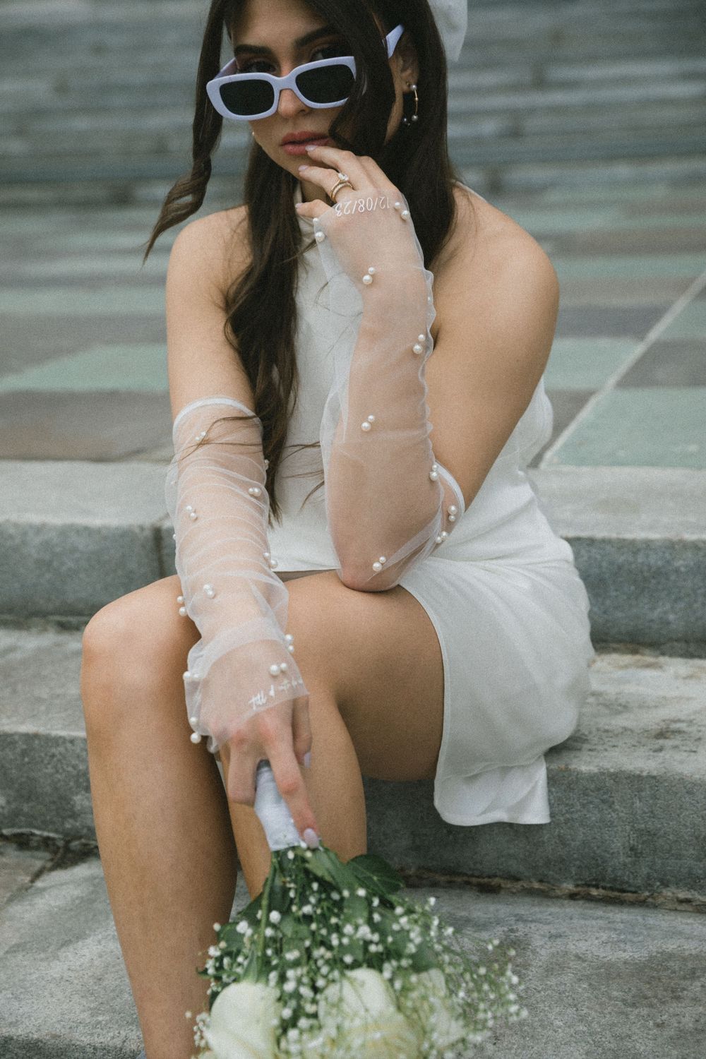 Bride in white mini dress, sheer pearl gloves, and lavender sunglasses sits on steps holding a white bouquet.