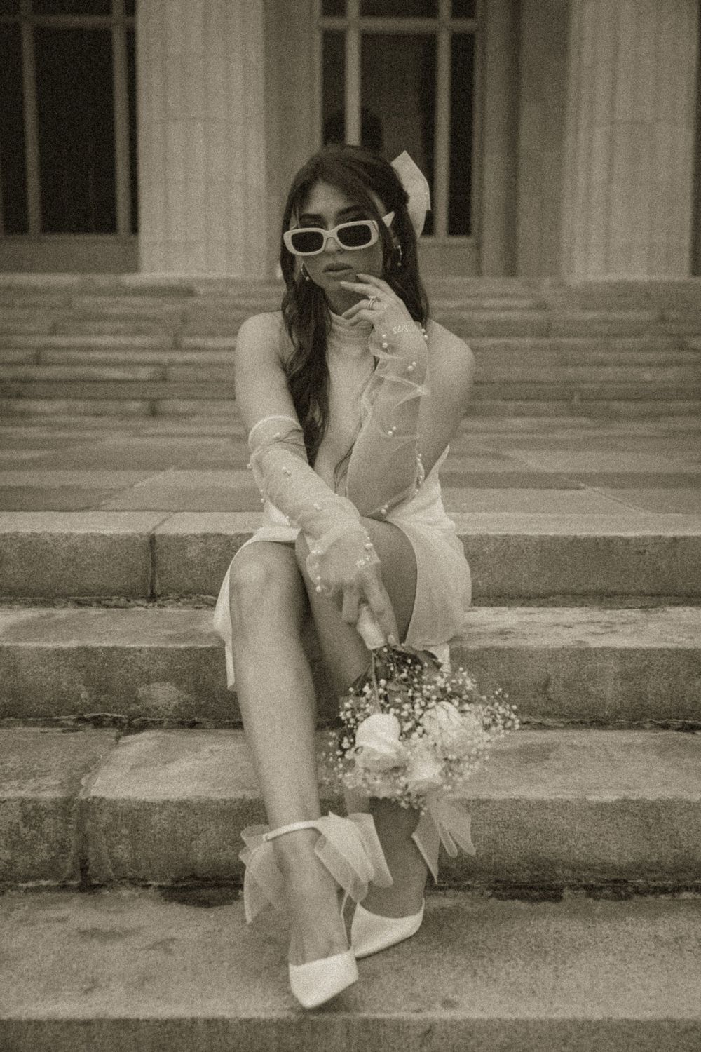 Bride in white dress, gloves, sunglasses sits on stone steps holding a small bouquet, vintage black and white photo.
