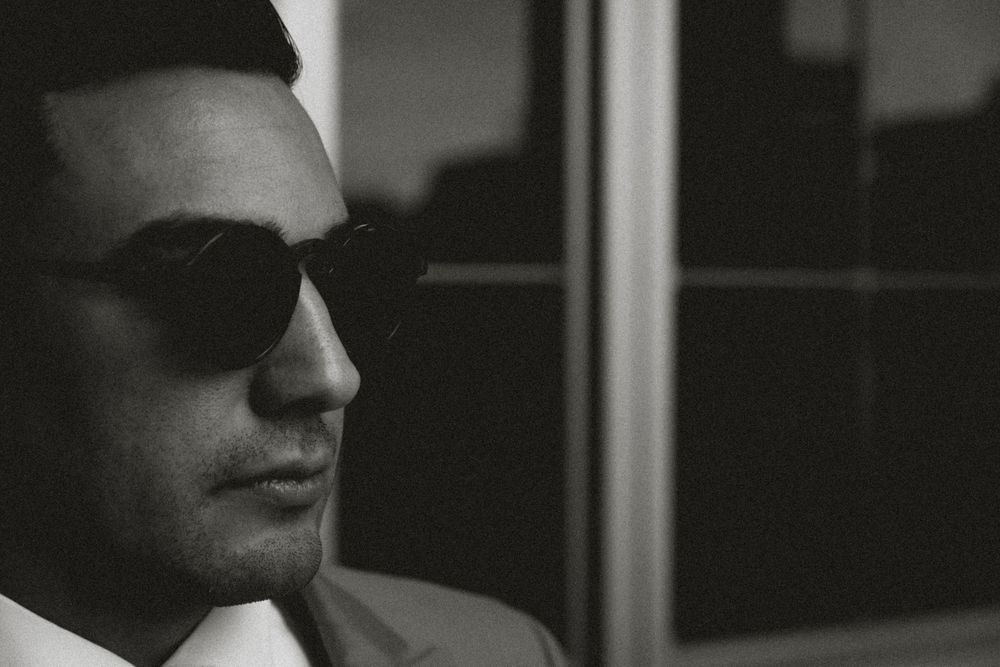 Black and white portrait of a man wearing sunglasses and a suit, looking away in a moody, cinematic style.
