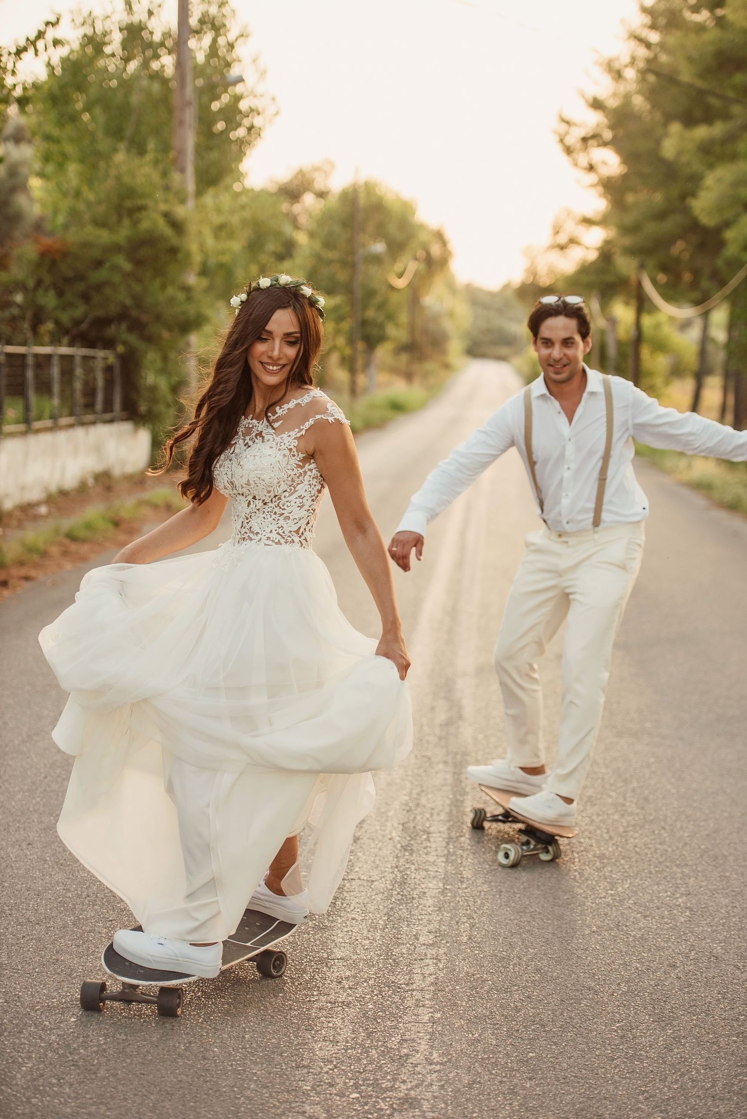 Bride in white lace gown and groom in suspenders skateboard down a sunny tree-lined road on their wedding day.