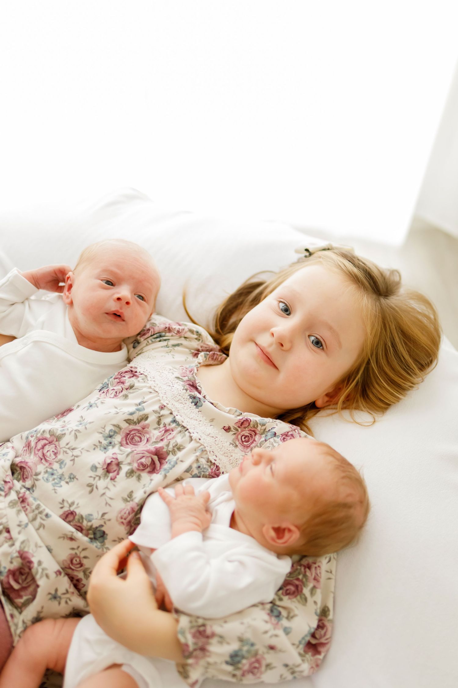 Young girl in floral dress lying on white bed with two newborn babies, sibling portrait photo.