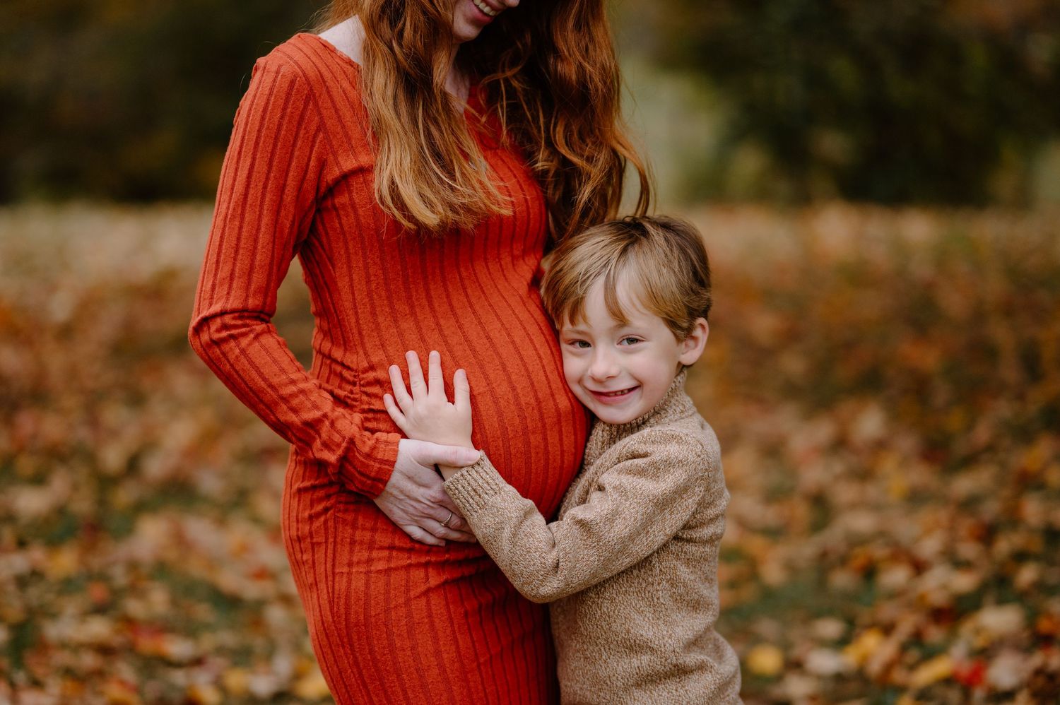 A young boy hugs his pregnant mother's belly outdoors in autumn, both wearing warm sweaters among fall foliage.
