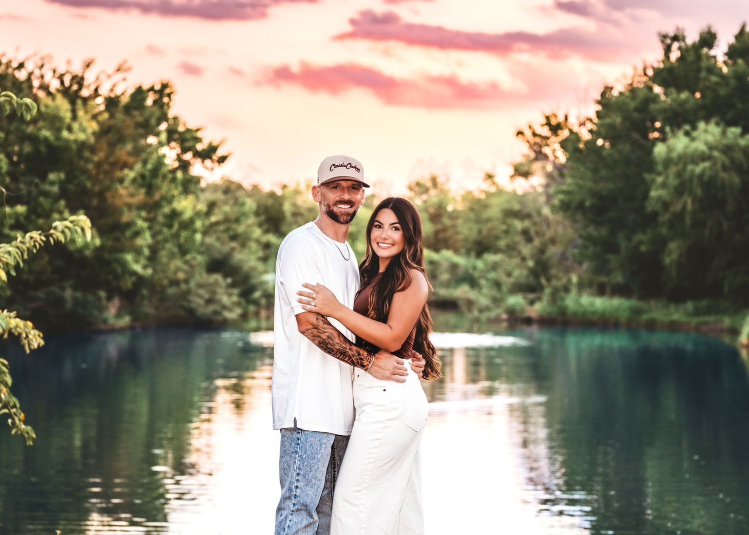 Couple embracing by a serene lake at sunset with lush green trees and a colorful pink sky in the background.