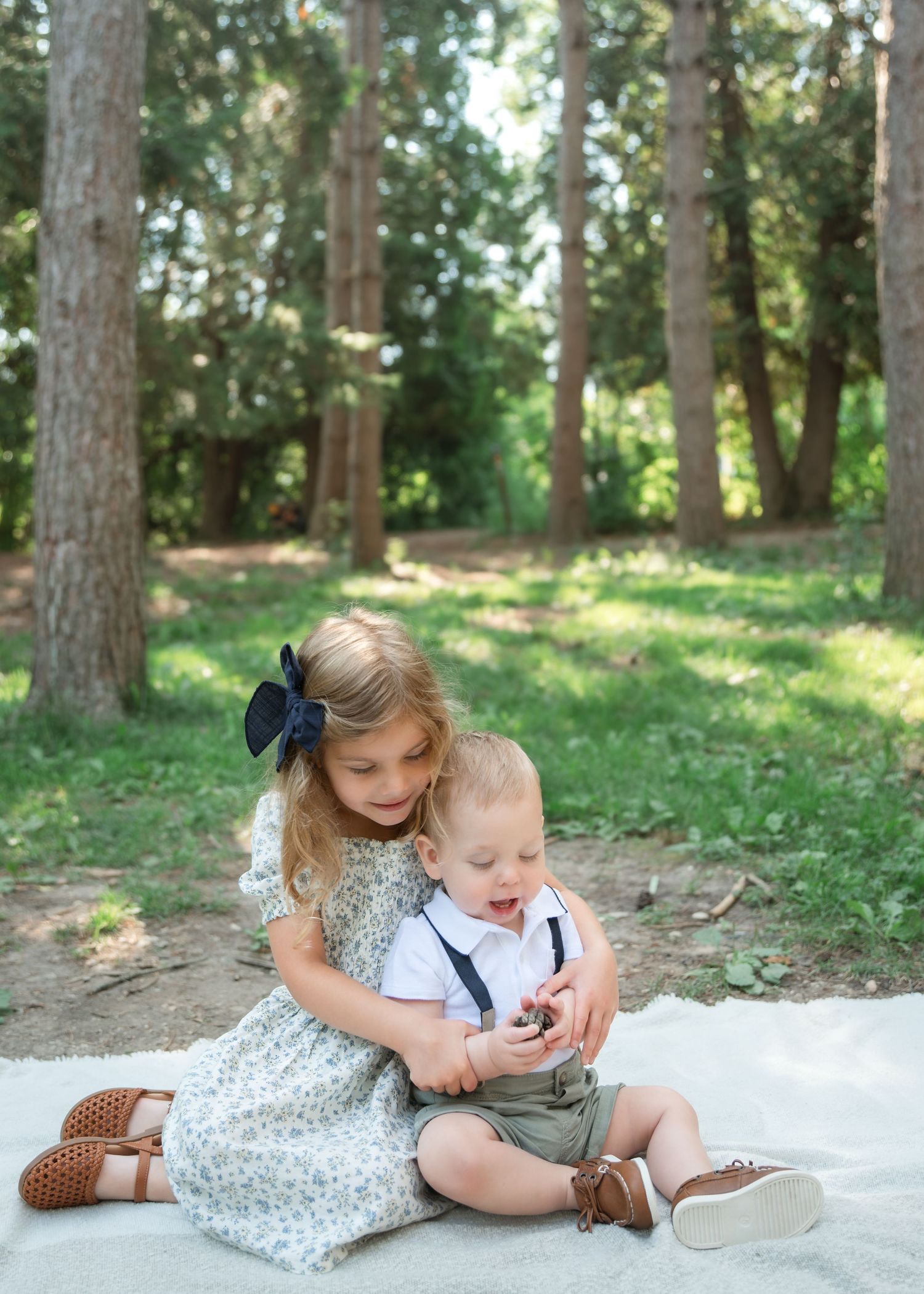 Two young siblings sit on a blanket in a forest, with an older girl in a floral dress hugging her baby brother.