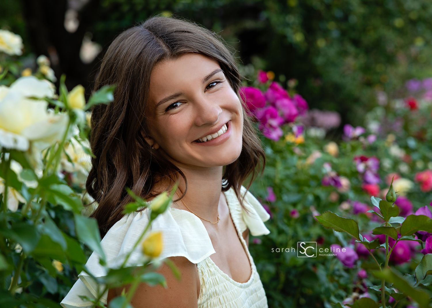 Young woman smiling among colorful garden roses, wearing white dress, outdoor senior portrait photography.