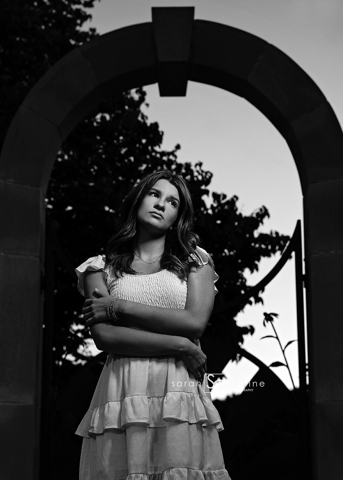 Black and white portrait of a young woman in a ruffled dress standing under a stone arch at dusk.