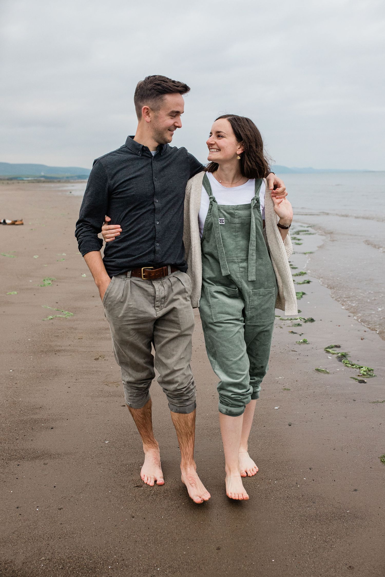 Smiling couple walks barefoot together along a sandy beach on a cloudy day.