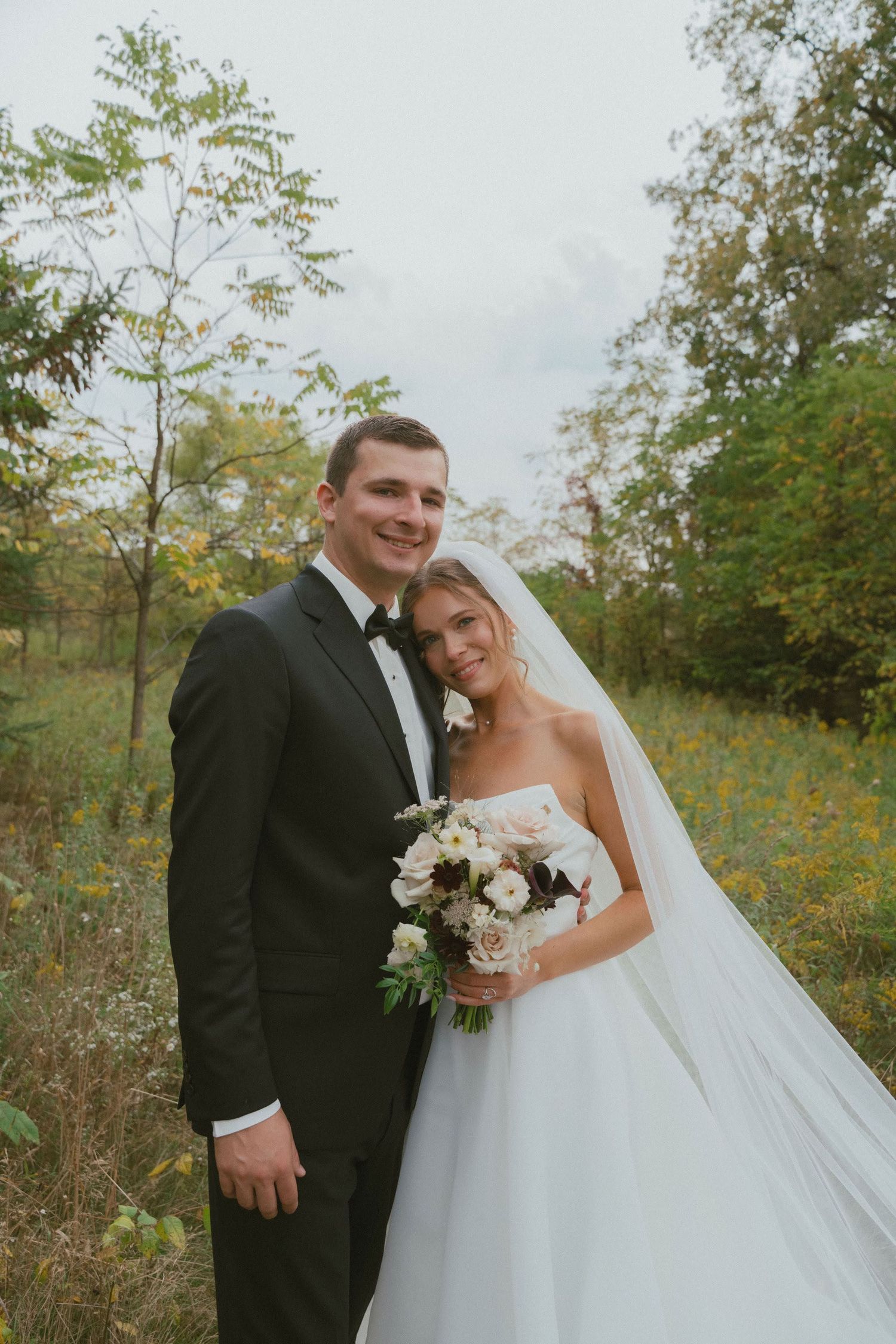 Bride in white gown holding elegant bouquet of blush roses, white flowers, and dark calla lilies beside groom in olive suit.