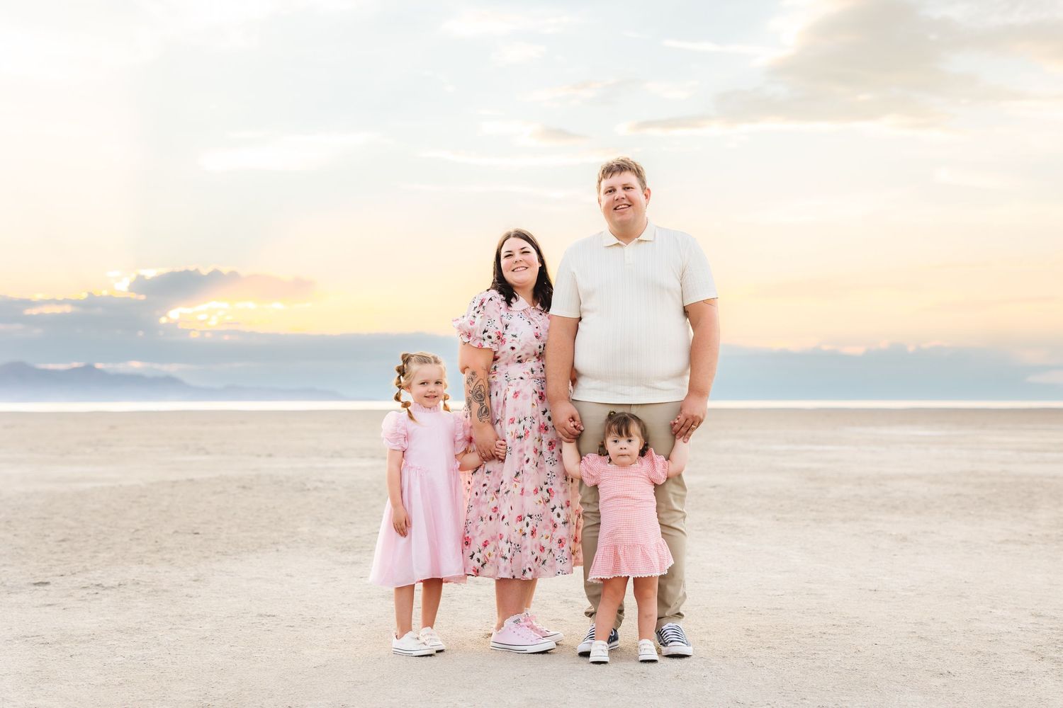 Family of four posing on a beach at sunset, parents holding hands with two young daughters in pink dresses. Utah Family Photographer