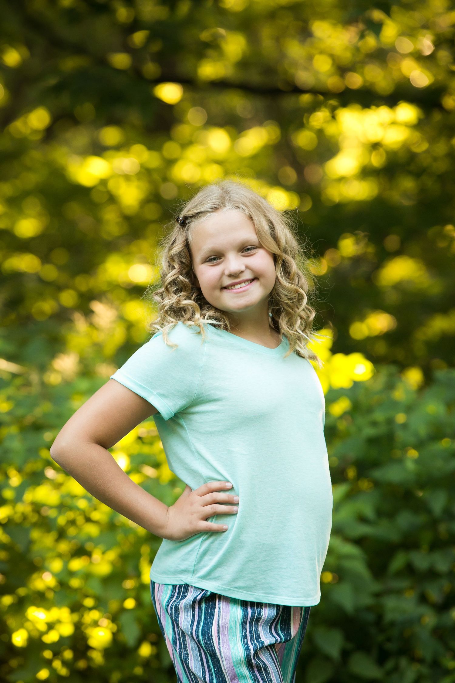 A smiling young girl with curly blonde hair wearing a mint green t-shirt poses outdoors with hands on hips.