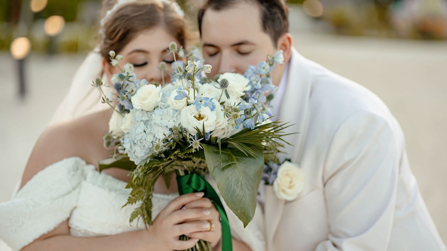 A bride and groom share a romantic moment smelling a blue and white wedding bouquet outdoors.