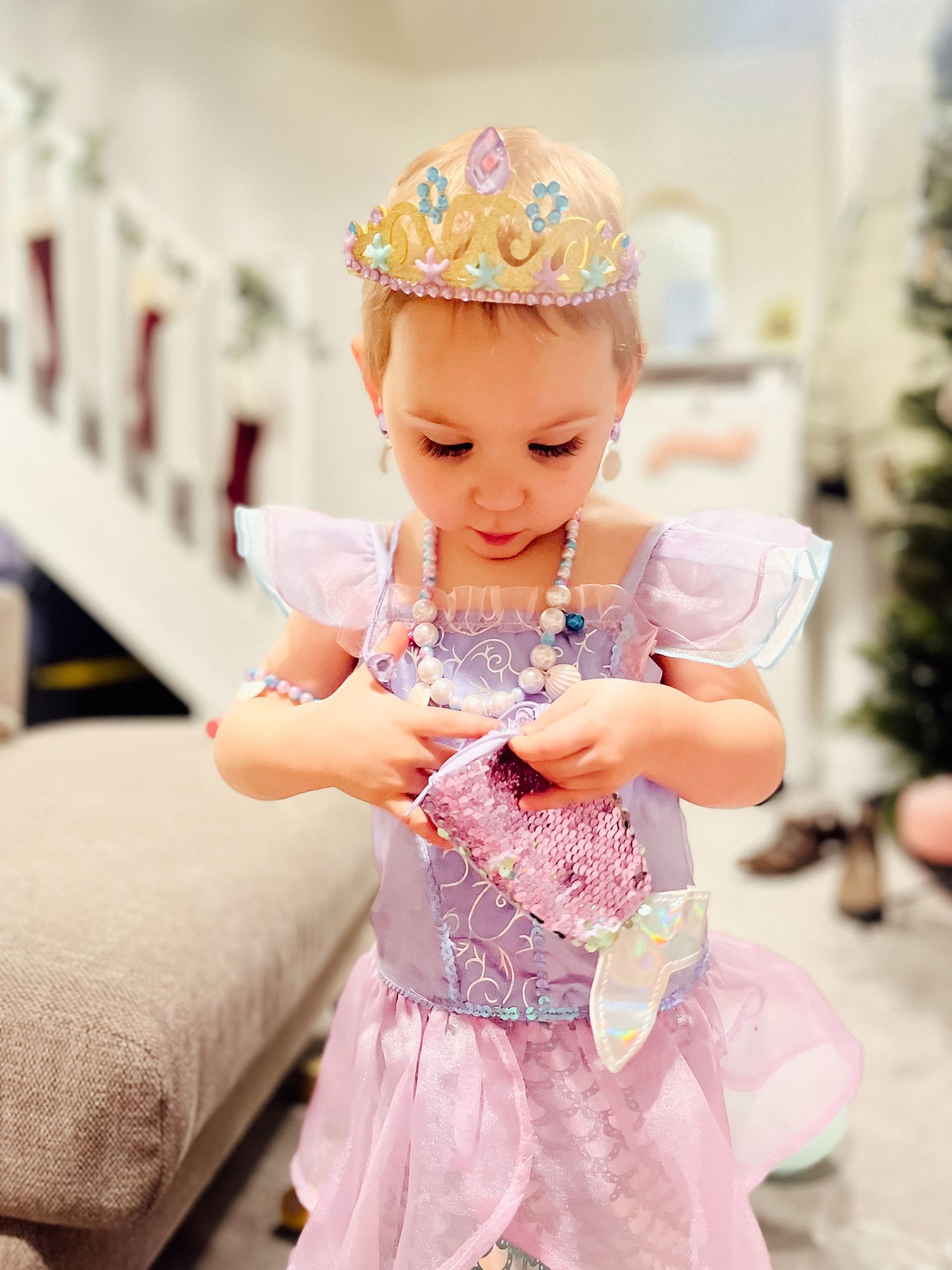 Young girl in pink princess dress and tiara opens a sparkly gift bag while playing dress-up indoors.