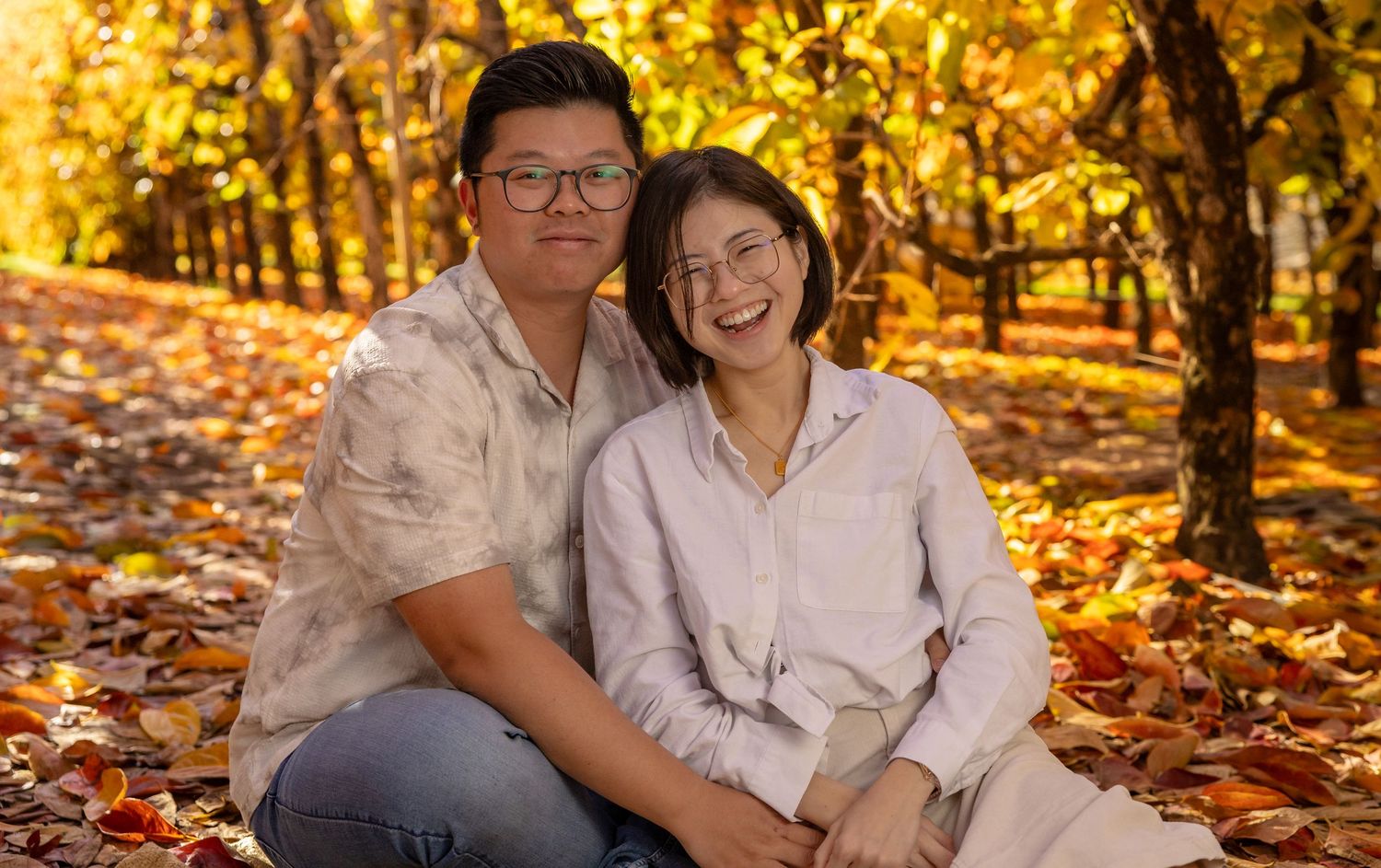 Couple smiling together among golden autumn leaves in a sunlit orchard during fall season.