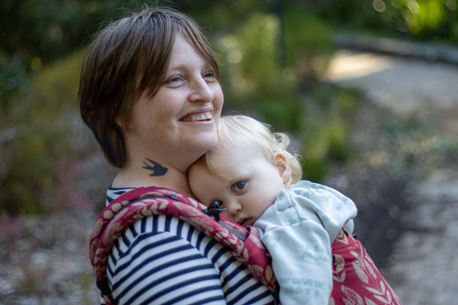 Woman carrying newborn baby in colorful rainbow dragon wrap sling carrier outdoors in natural setting.