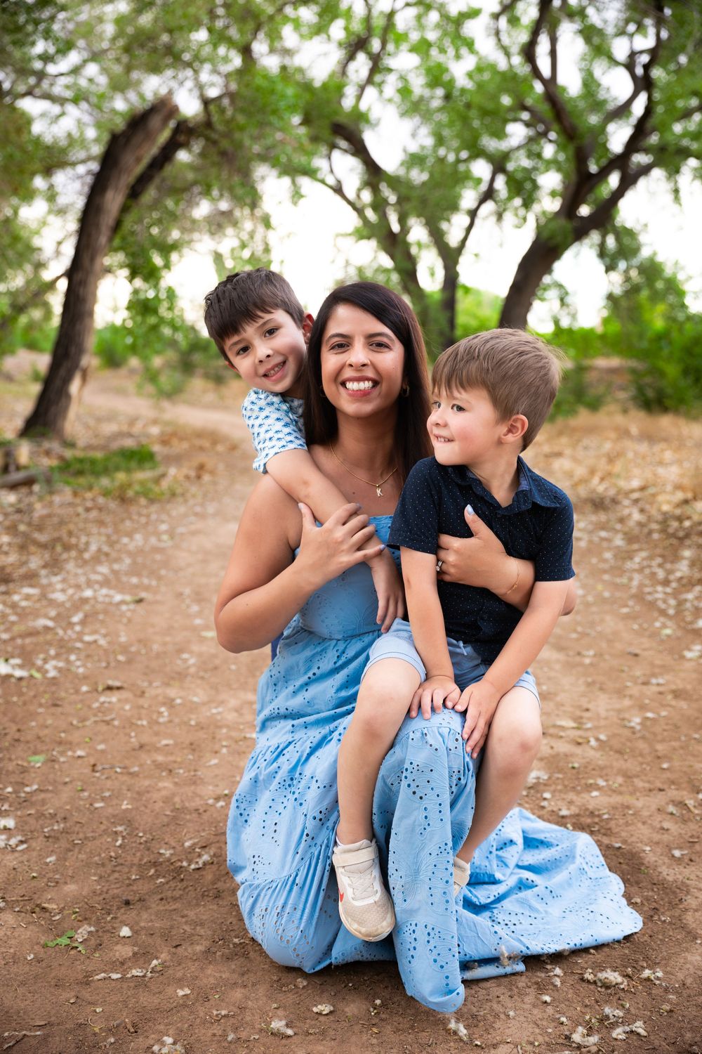 A smiling woman in a blue dress sits outdoors with two young boys hugging her, surrounded by trees.