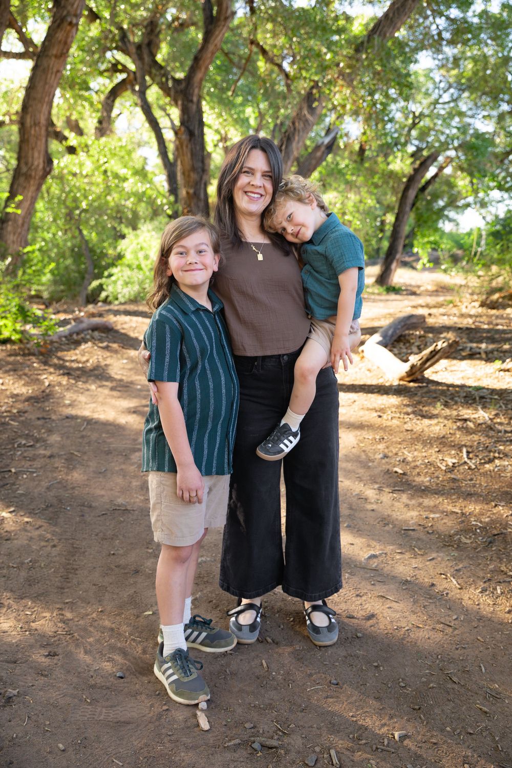 A woman smiles holding a young child while standing with another child on a wooded trail outdoors.