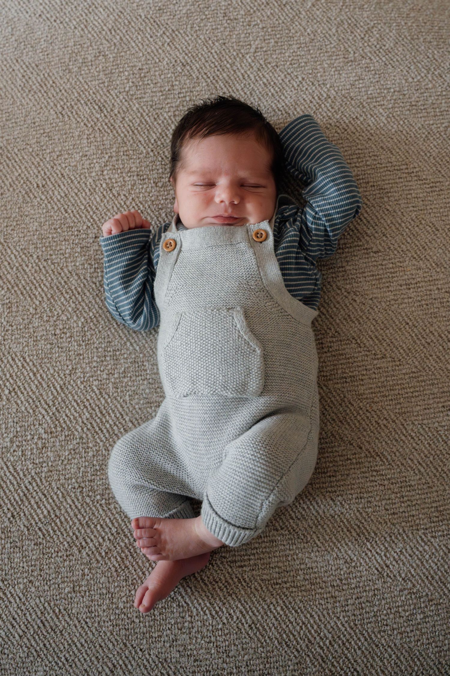 Newborn baby sleeping peacefully in gray knit overalls and striped shirt on carpet.