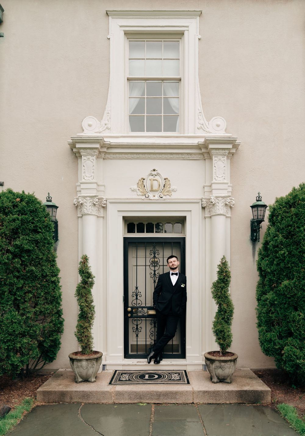 Groom in black tuxedo and bow tie poses elegantly at ornate white doorway of classic estate building.