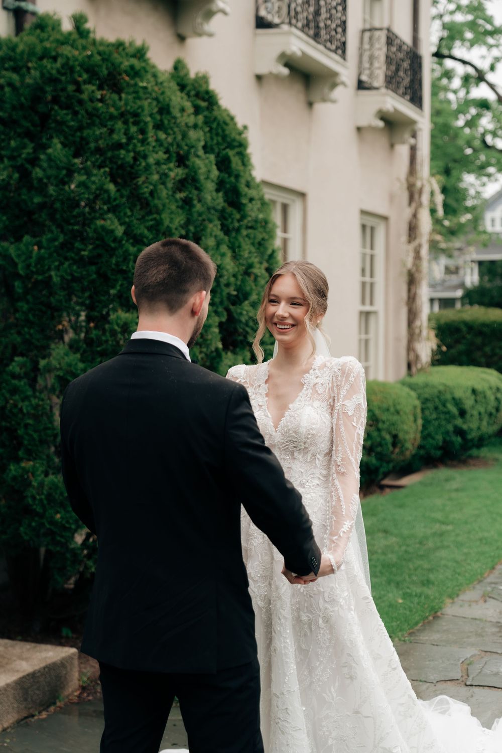 Smiling bride in long-sleeve lace gown holds hands with tuxedo-clad groom outside elegant manor estate.
