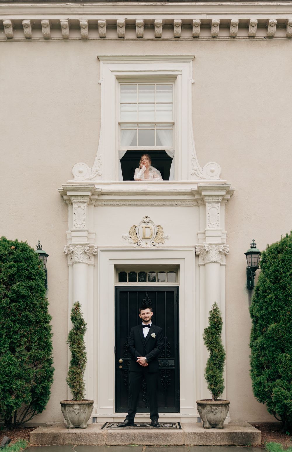 Groom in tuxedo stands at mansion door while bride in wedding dress looks down from upper window above.