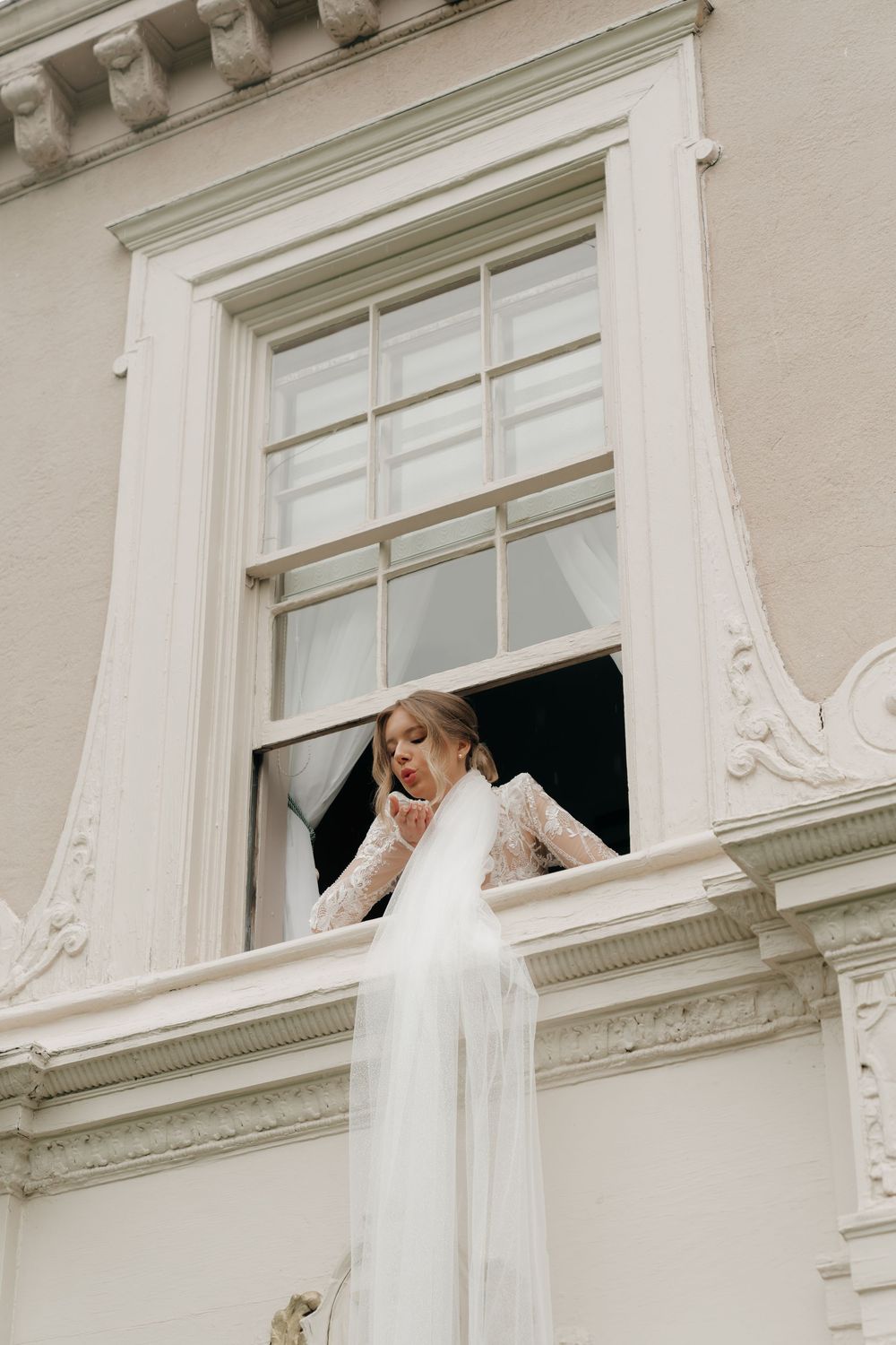 Bride in lace wedding dress and veil leaning out of ornate white building window.