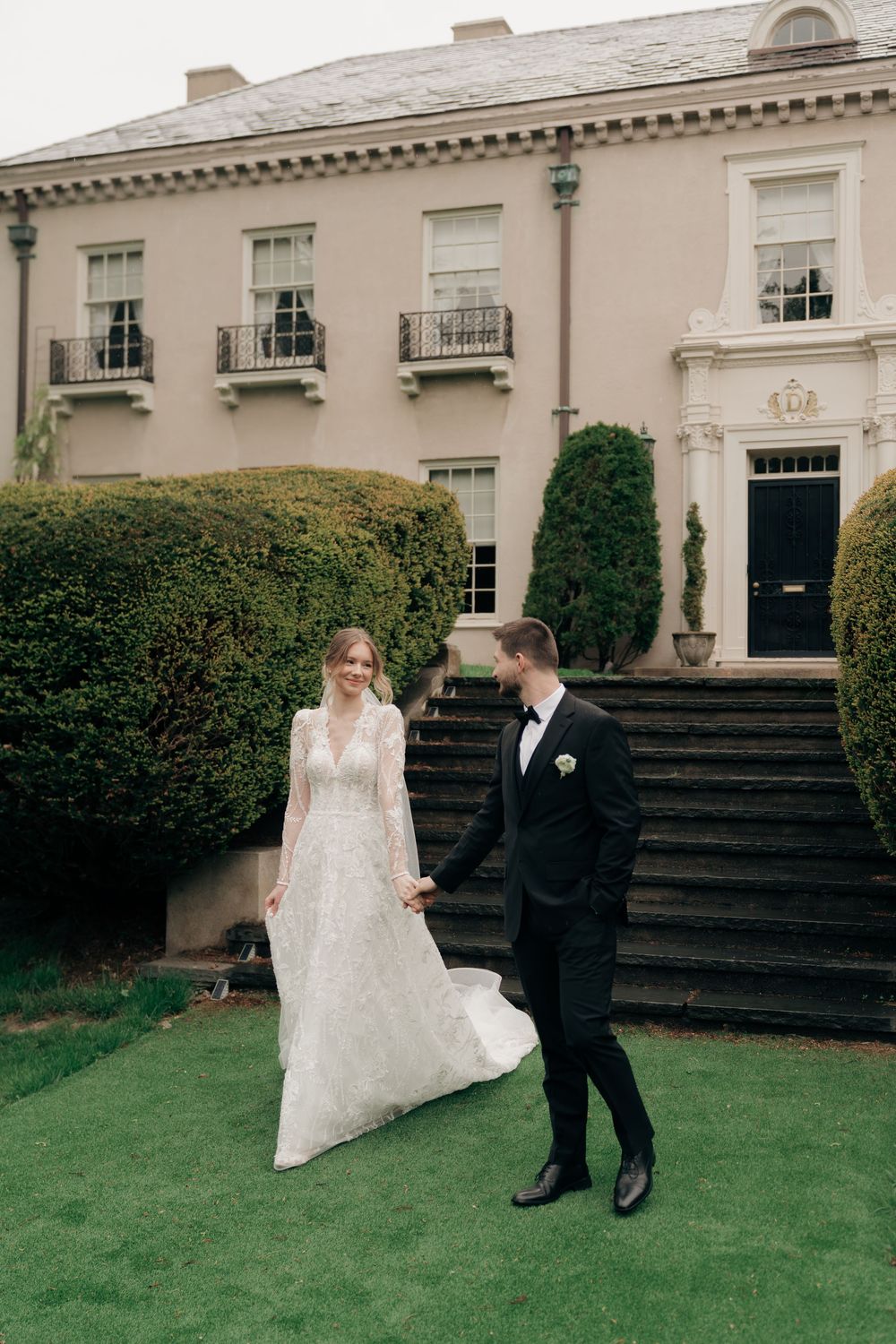 Bride and groom hold hands on lawn before grand estate in lace dress and black tuxedo.