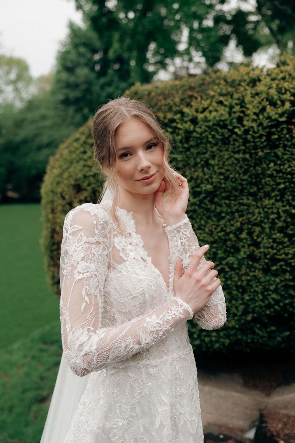 A bride in a long-sleeve lace wedding gown poses gracefully outdoors near trimmed hedges and lush greenery.