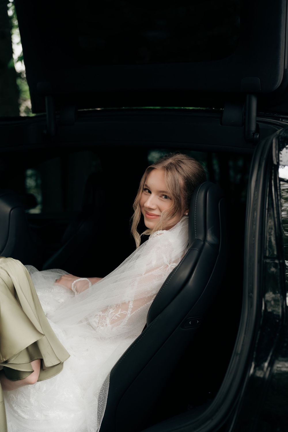 Beautiful bride in lace wedding dress sitting in luxury car backseat smiling softly.