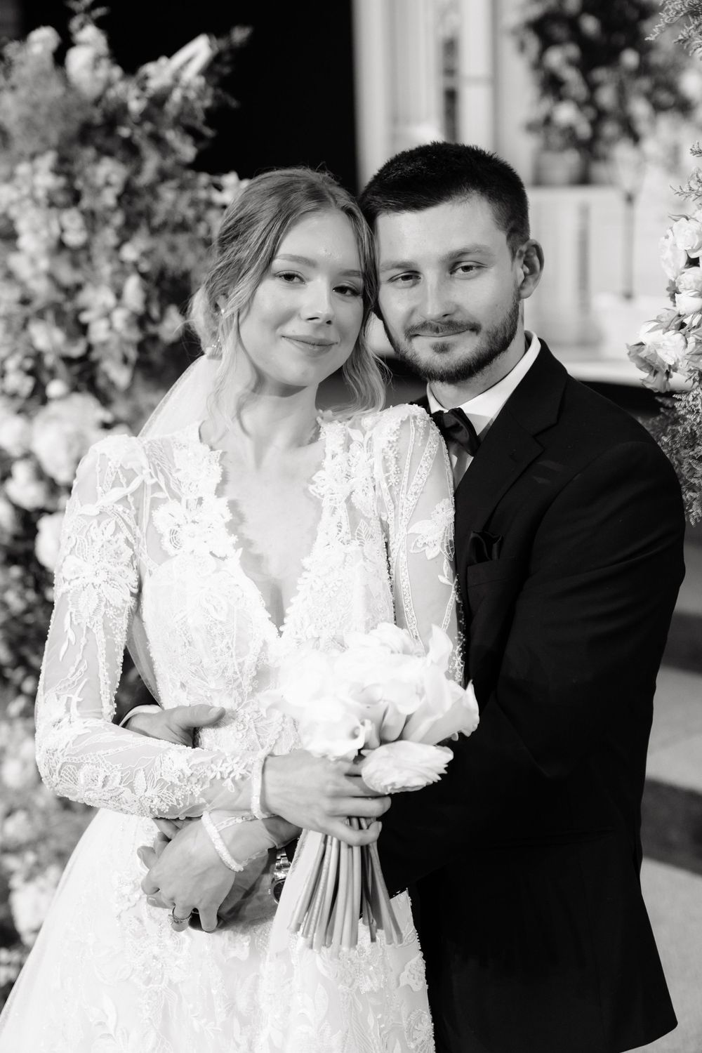 Newlywed couple posing with white bouquet in black and white portrait at floral ceremony arch.