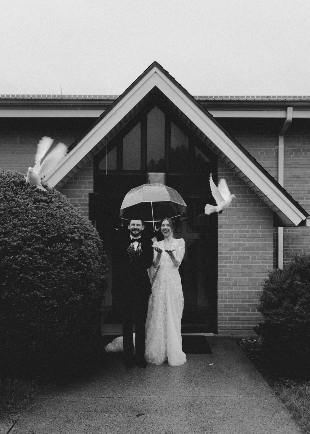 Newlyweds releasing white doves under umbrella outside church entrance in black and white photo.
